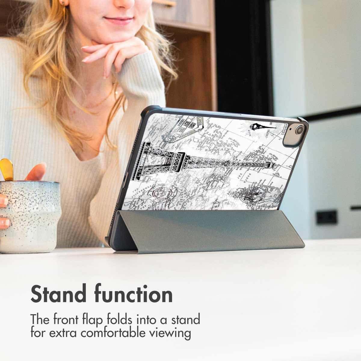 Een tablet in een grijze hoes met Eiffeltoren-kaartontwerp staat op een tafel naast een vrouw met een mok.