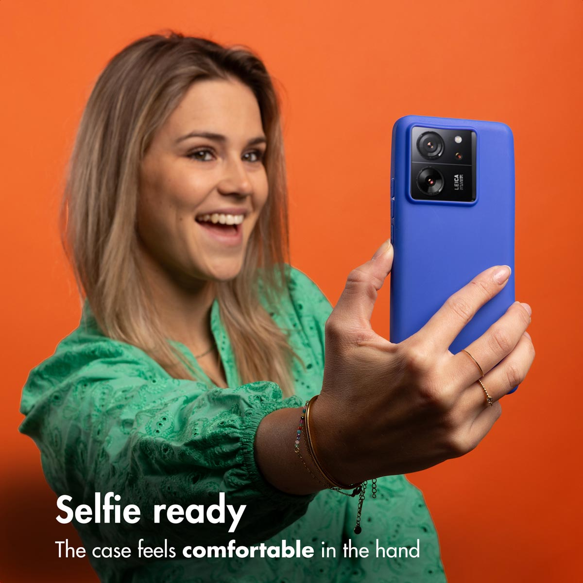 Una mujer rubia con camisa verde se hace un selfie con un smartphone azul y funda.