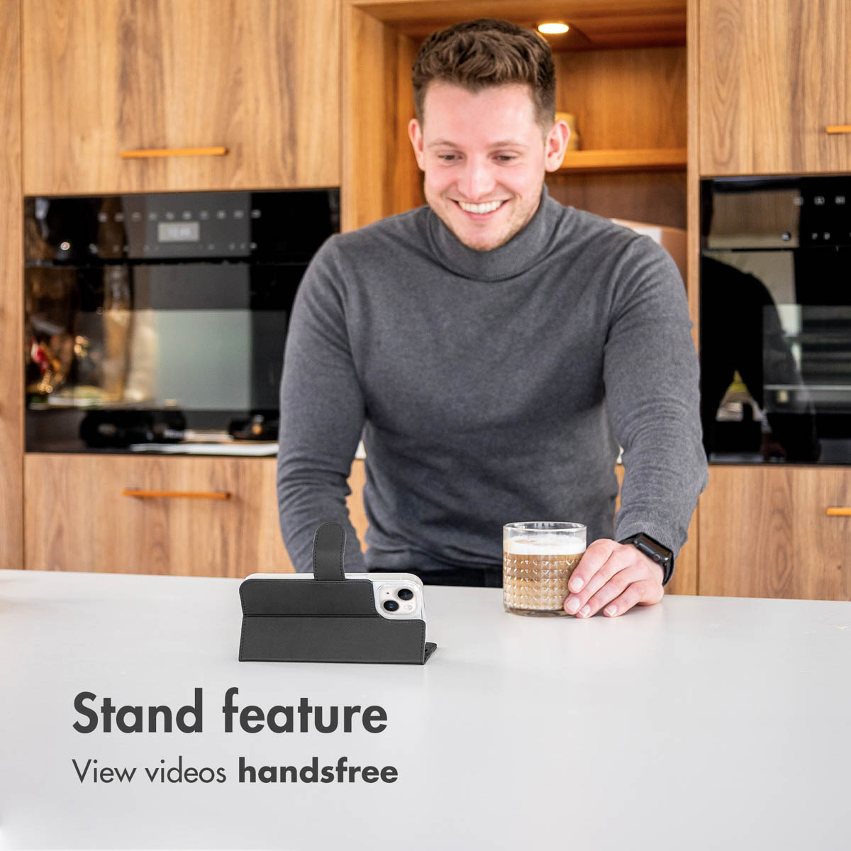 Un hombre en una cocina sonríe a un teléfono inteligente en una funda con soporte al lado de un vaso.
