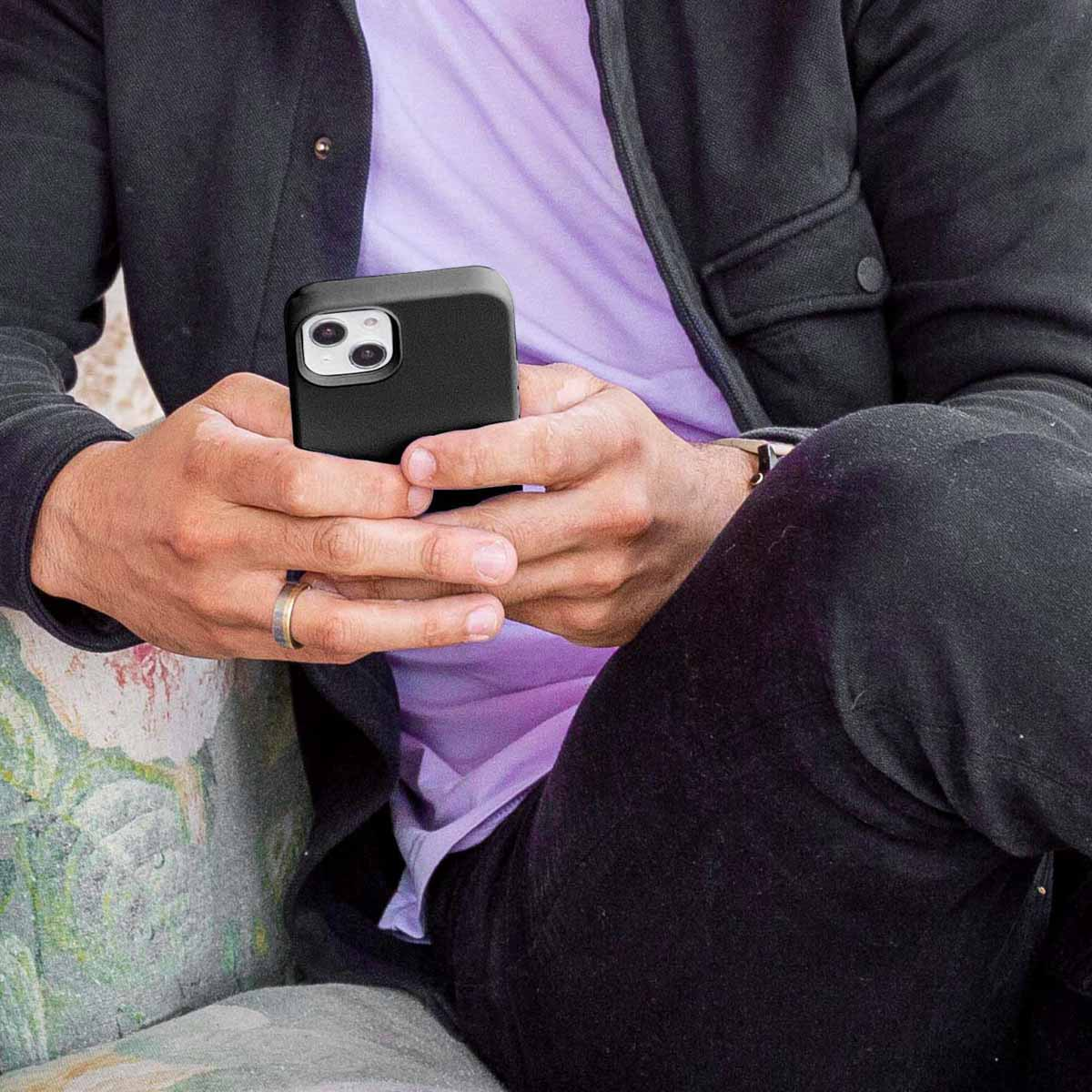 Siedząca osoba trzyma czarny smartfon, ubrana w ciemną kurtkę, jasnofioletową koszulkę i ciemne spodnie.