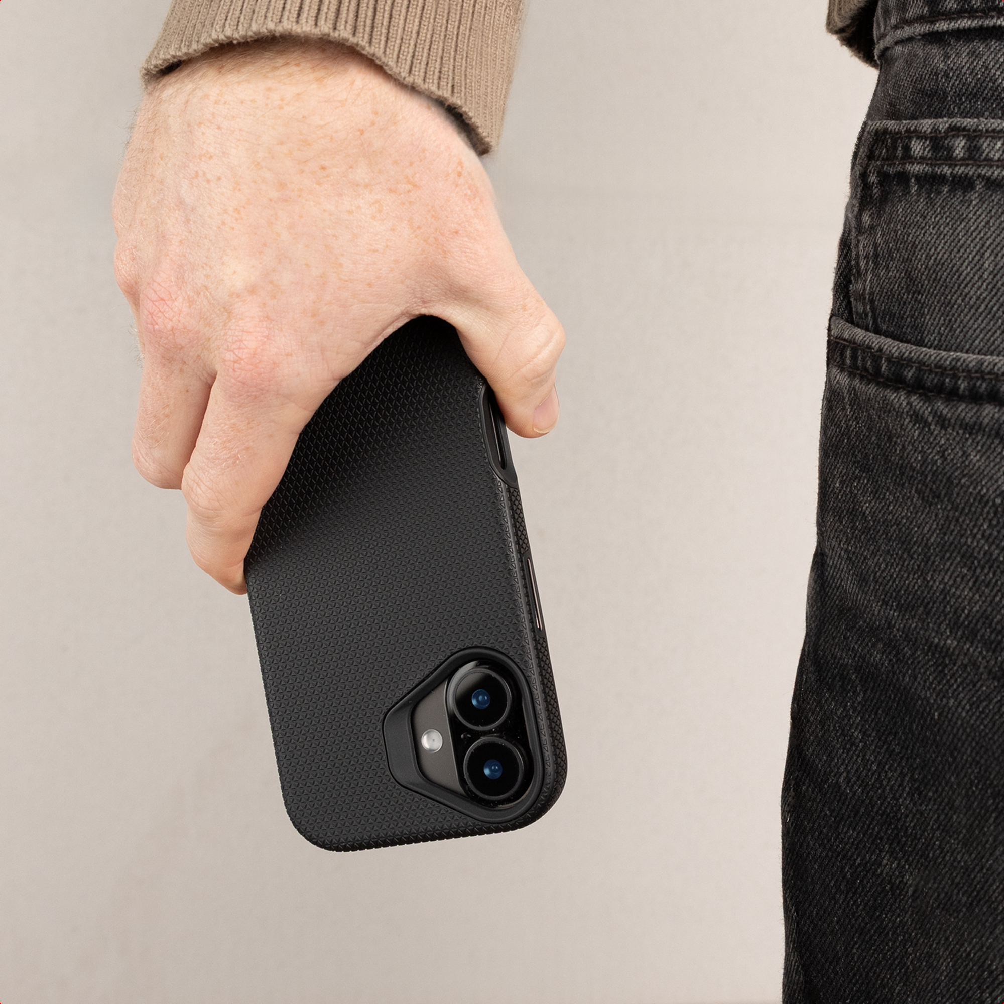 Una mano tiene uno smartphone nero in una custodia protettiva con doppia fotocamera su sfondo chiaro.