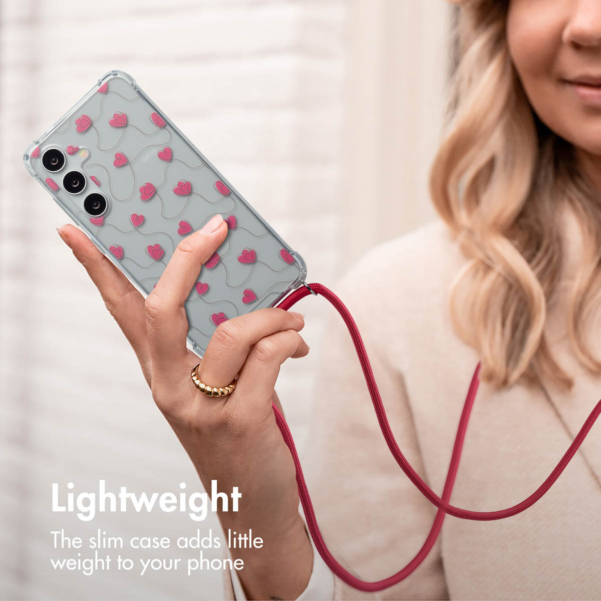 Une femme blonde tient un smartphone dans une coque transparente avec des cœurs roses et une lanière rouge.