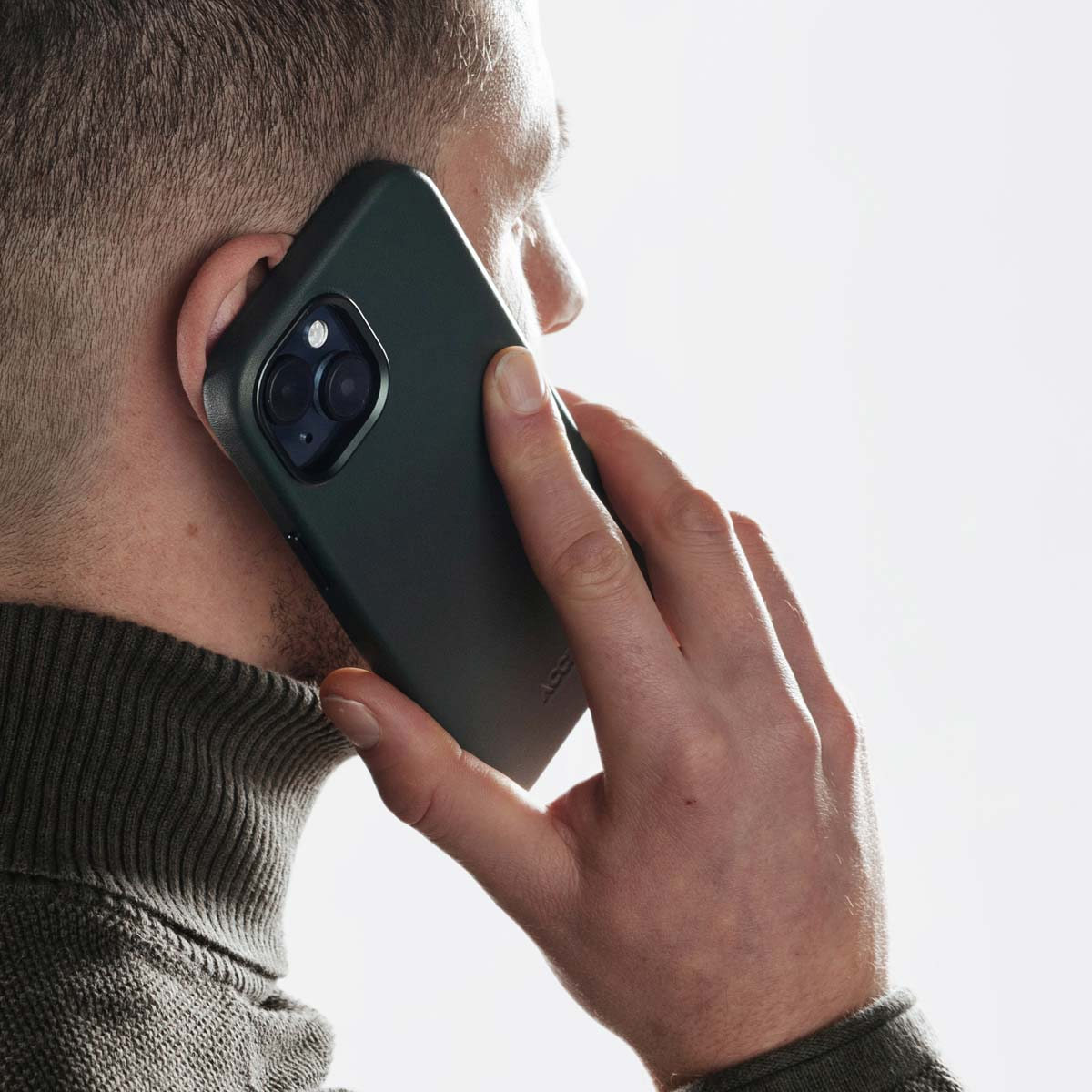Un uomo con un dolcevita verde tiene uno smartphone verde scuro all'orecchio su sfondo bianco.