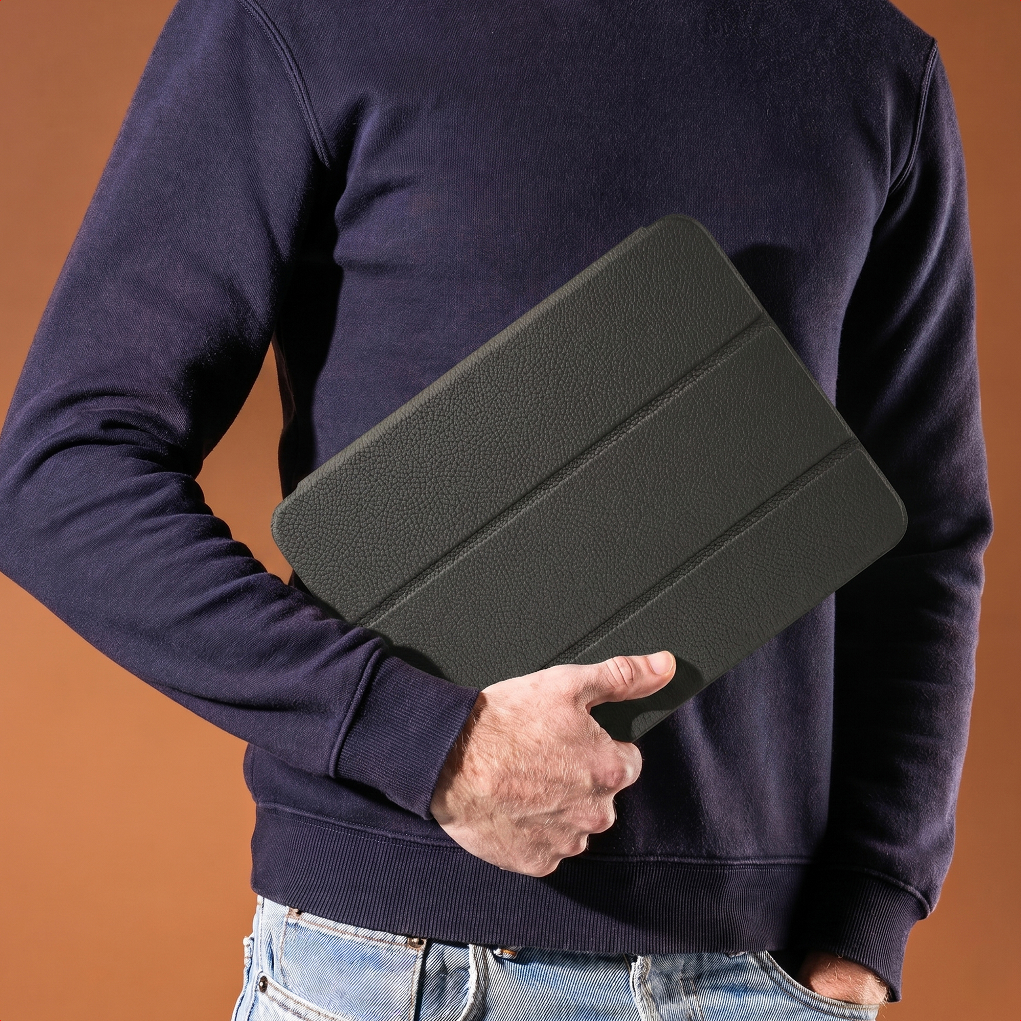 Iemand in donkere trui en jeans houdt een zwarte leren tablethoes vast tegen een bruine achtergrond.