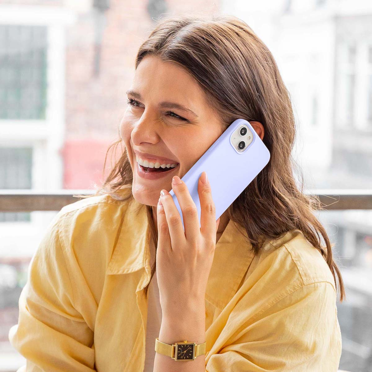 Een glimlachende vrouw in een geel shirt belt met een lichte paarse smartphone tegen een wazige achtergrond.