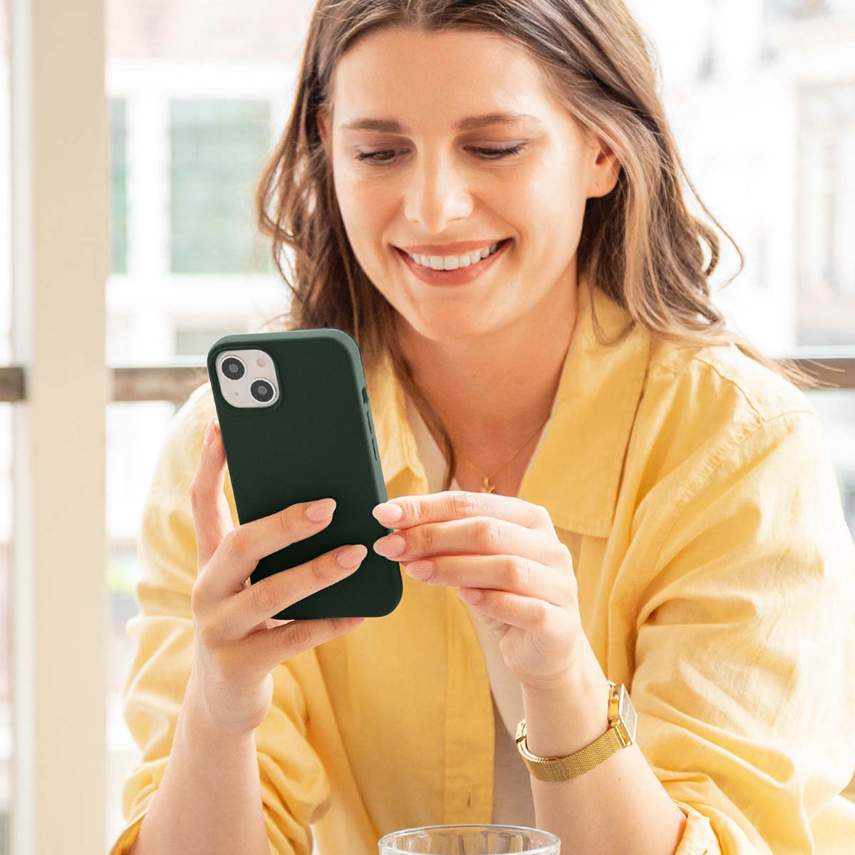 Uśmiechnięta kobieta w żółtej koszuli patrzy na swój ciemnozielony smartfon.