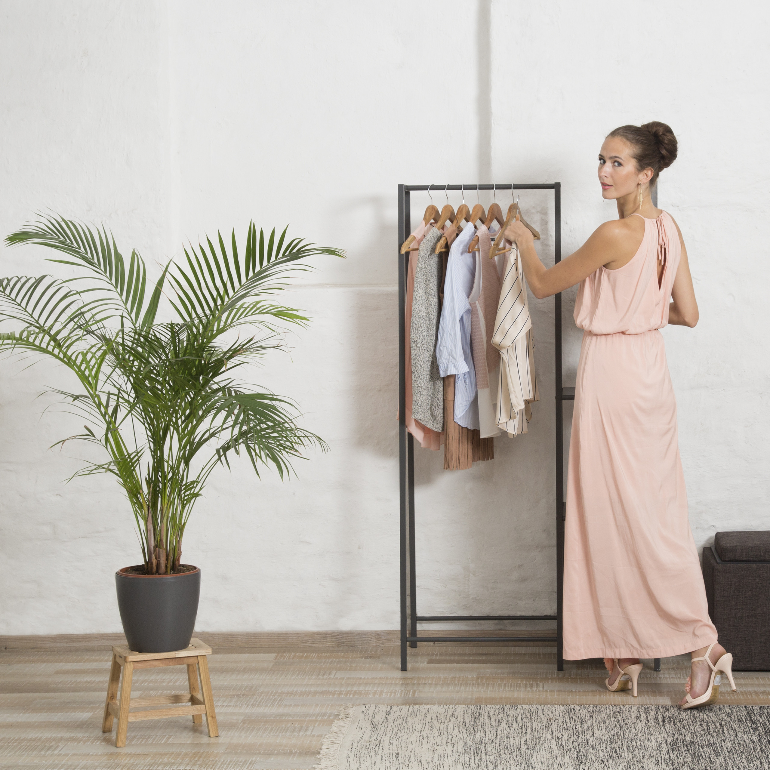 Eine Frau in einem langen rosa Kleid greift vor einer Kleiderstange und einer Zimmerpflanze nach Kleidung.