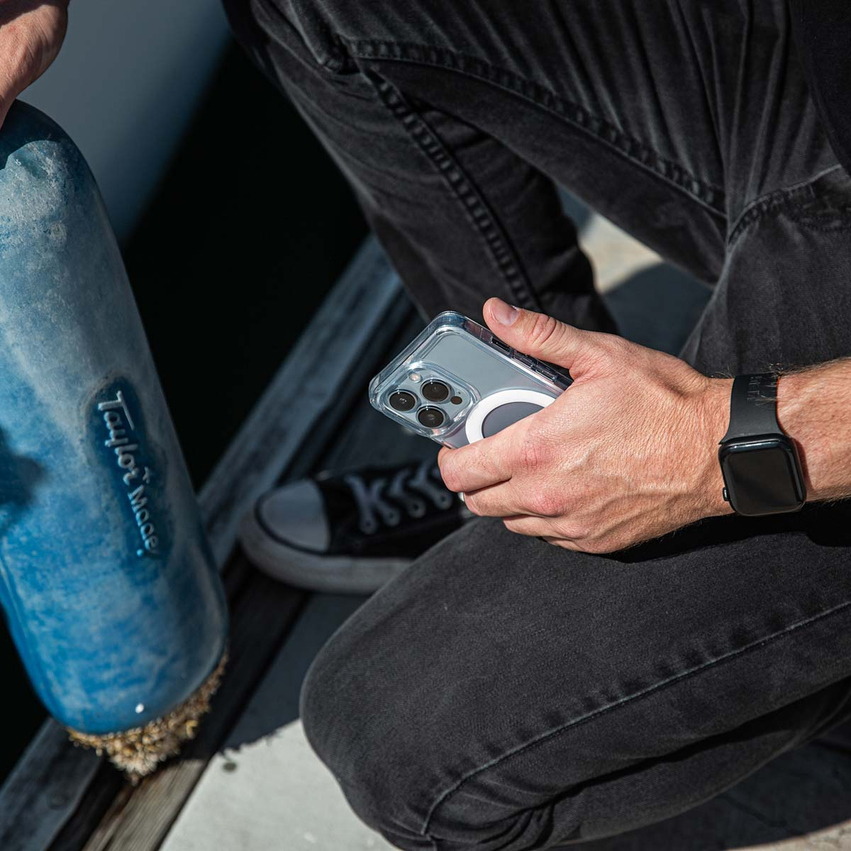 Person in dunklen Jeans hält ein Smartphone in durchsichtiger Hülle mit MagSafe-Ring und trägt eine schwarze Smartwatch.
