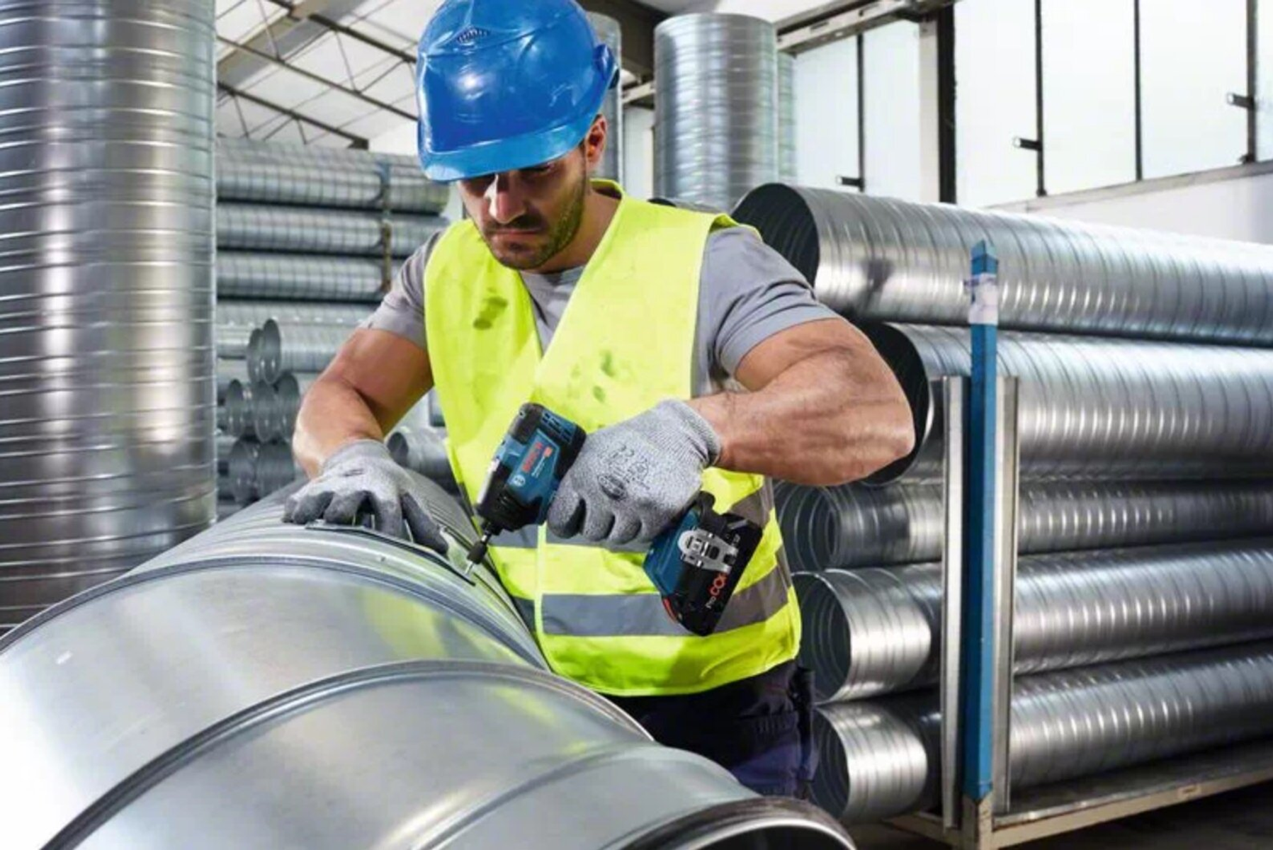 Ein Arbeiter mit Helm und Weste bohrt Löcher in ein großes Metallrohr in einer Fabrik.