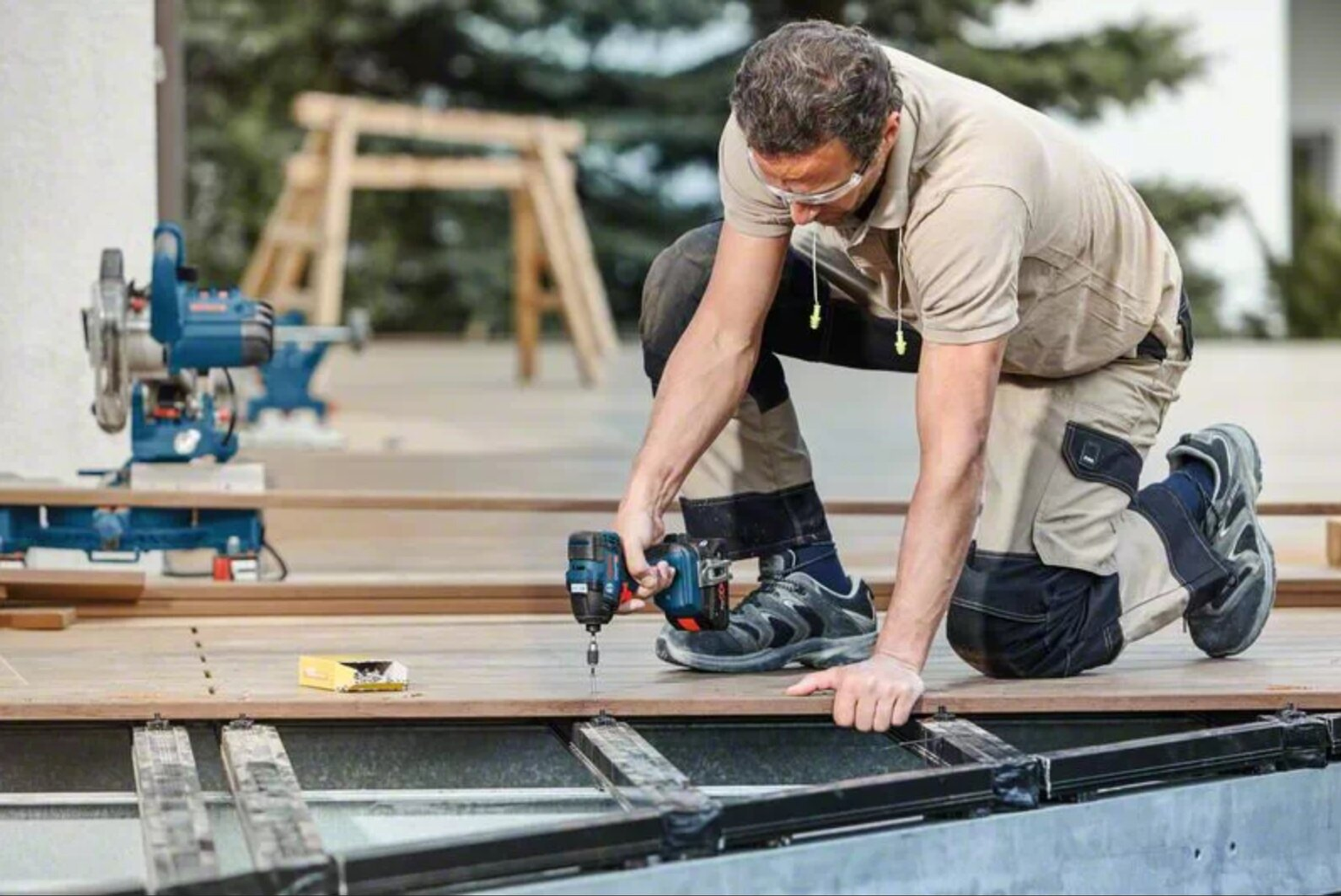 Ein Handwerker kniet auf einer Holzterrasse und verschraubt Dielen mit einem Akkuschrauber.