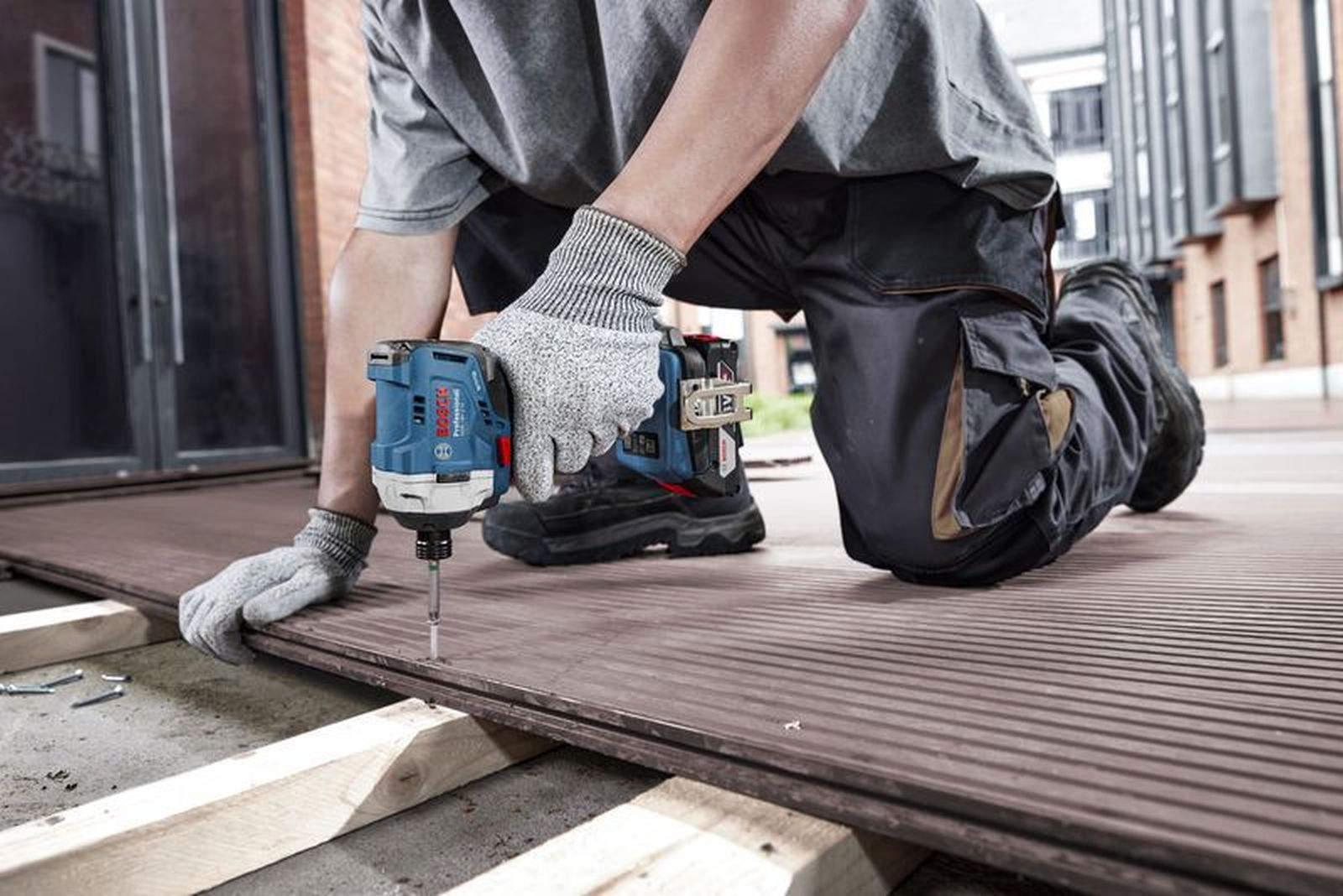Ein Arbeiter kniet auf einer Holzterrasse und verschraubt Dielen mit einem blauen Akkuschrauber.