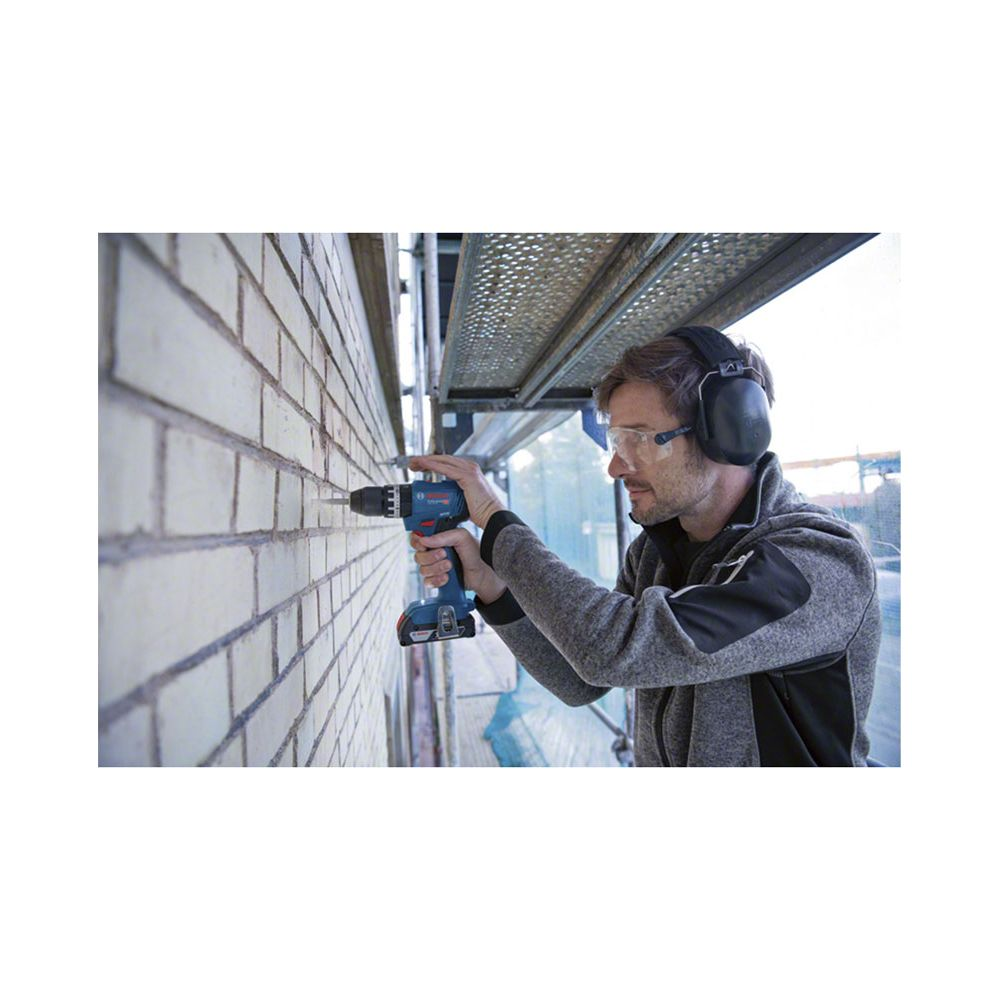 Un ouvrier utilise une perceuse bleue sur un mur de briques avec des lunettes de protection et un casque.