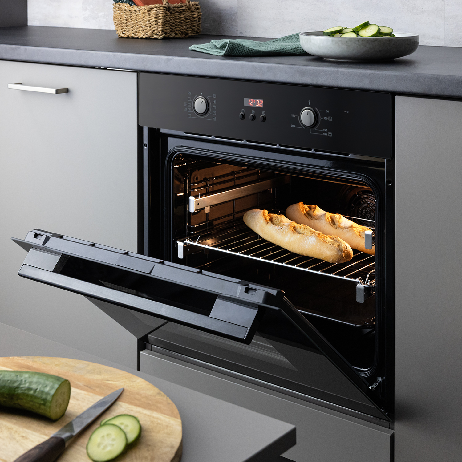 Ein offener, schwarzer Backofen mit zwei Baguettes auf dem Rost. Auf der Arbeitsfläche liegt Gemüse.