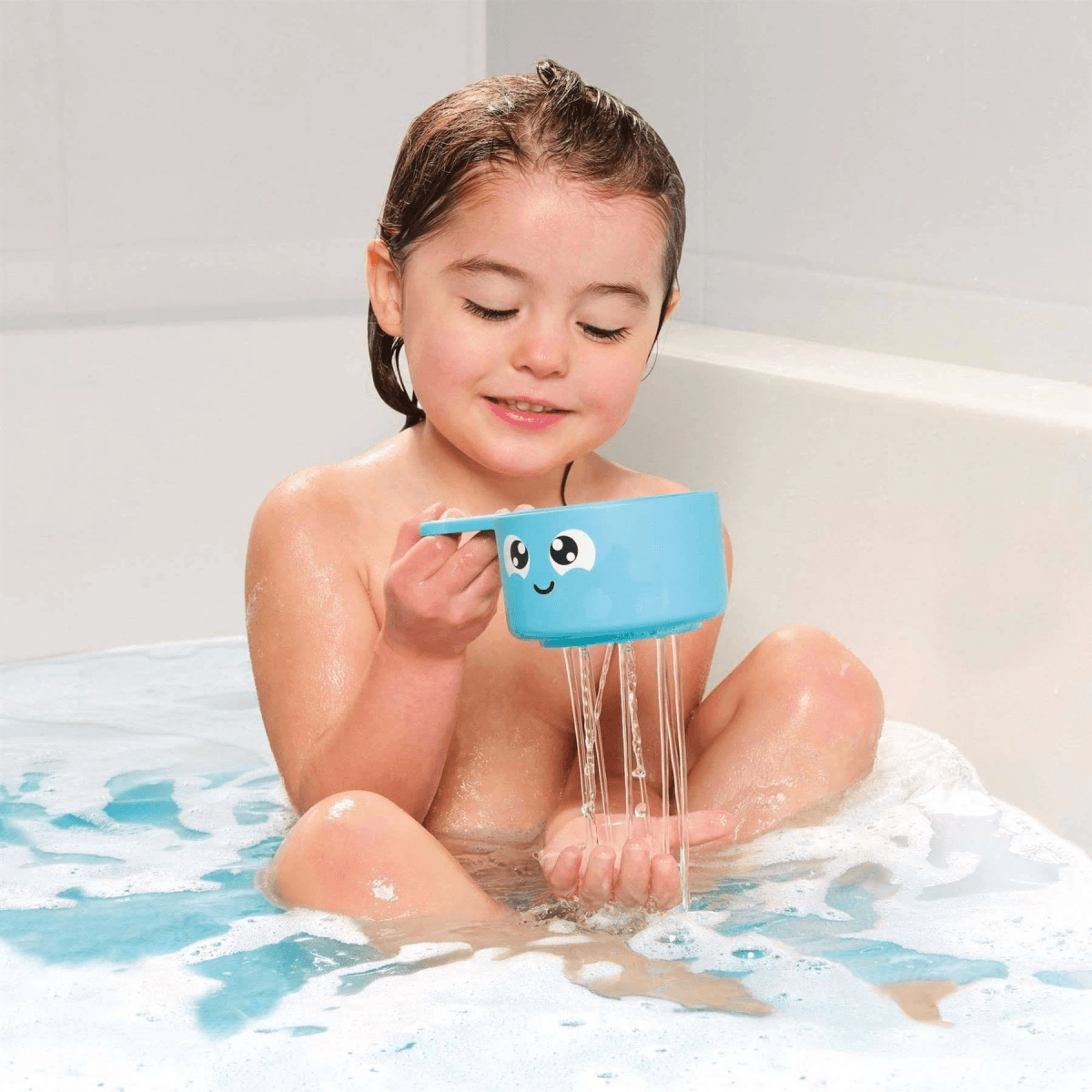Un niño se sienta en una bañera y vierte agua sobre sus manos desde una taza azul con cara.