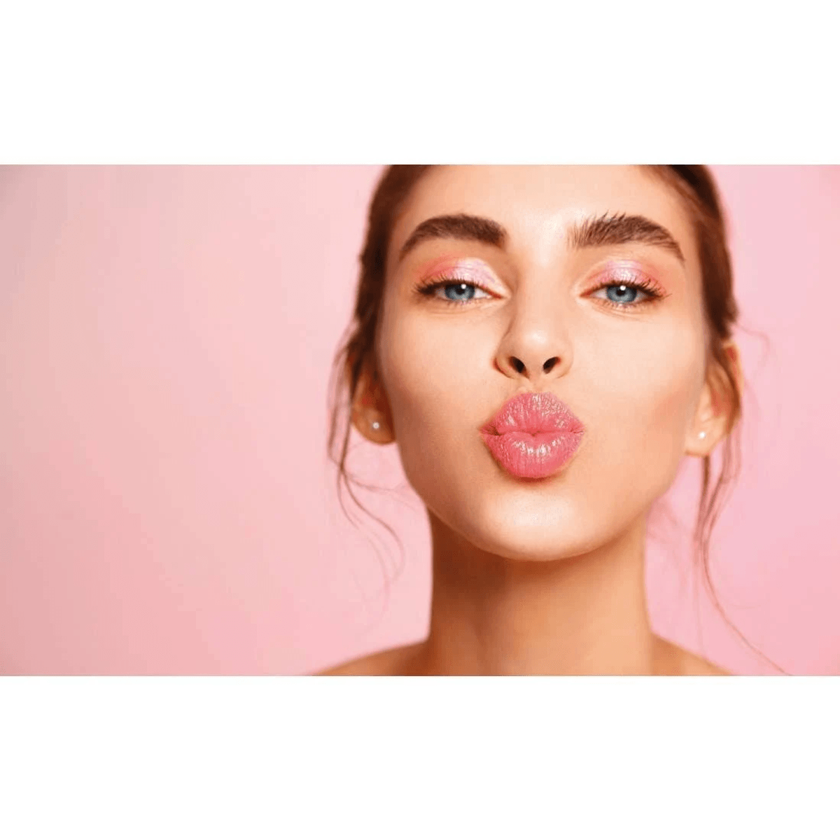 Primer plano de una mujer con ojos azules, pintalabios y sombra de ojos rosados, haciendo pucheros ante un fondo rosa.
