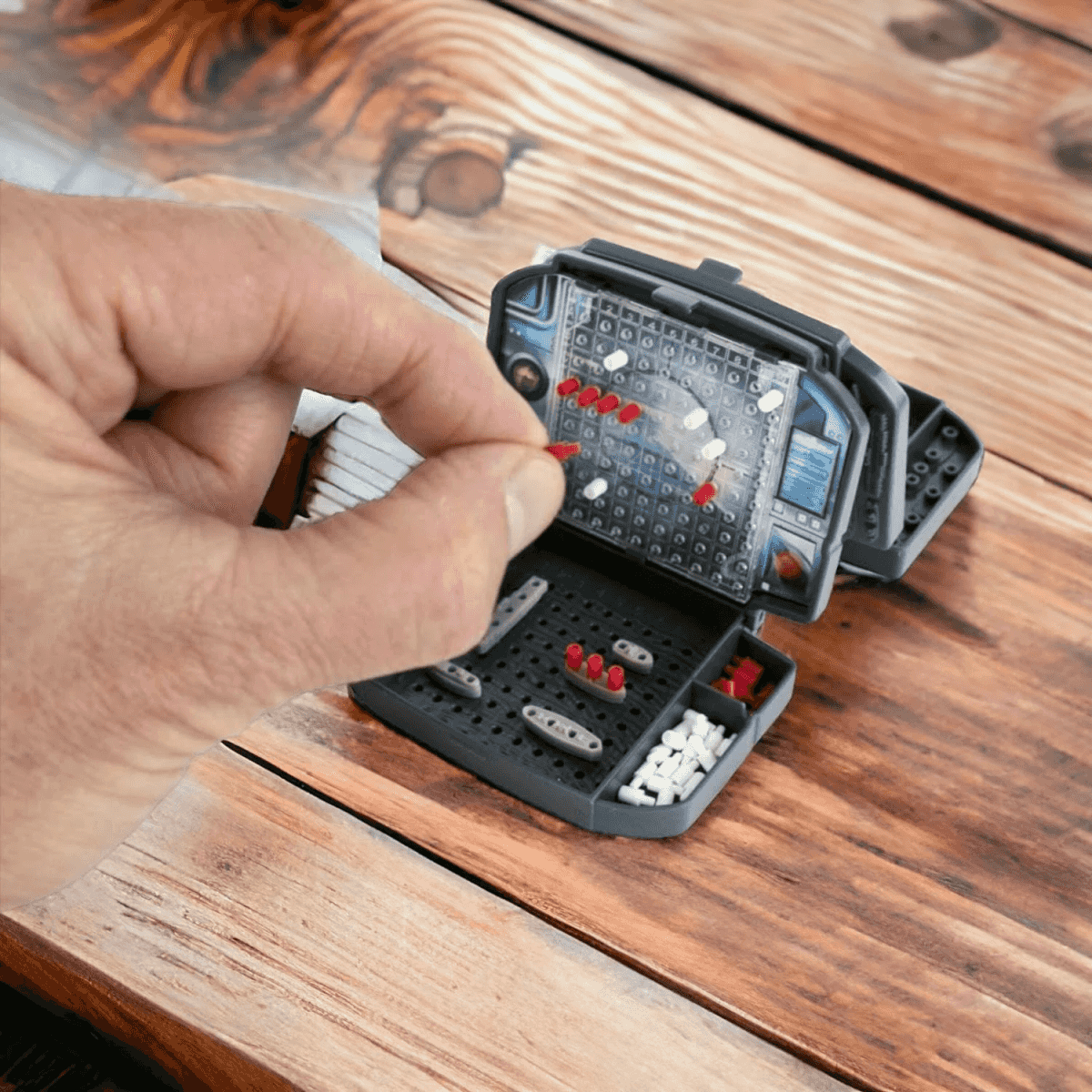 Una mano coloca una clavija roja en un juego de mesa de batalla naval gris sobre una mesa de madera.