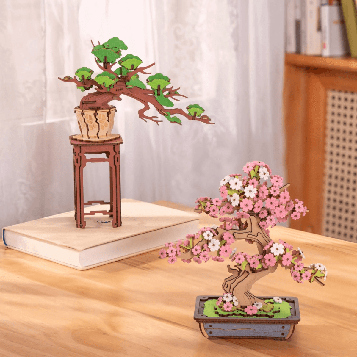 Dos modelos de bonsái de madera en una mesa: uno con hojas verdes en un soporte, el otro con flores rosas.