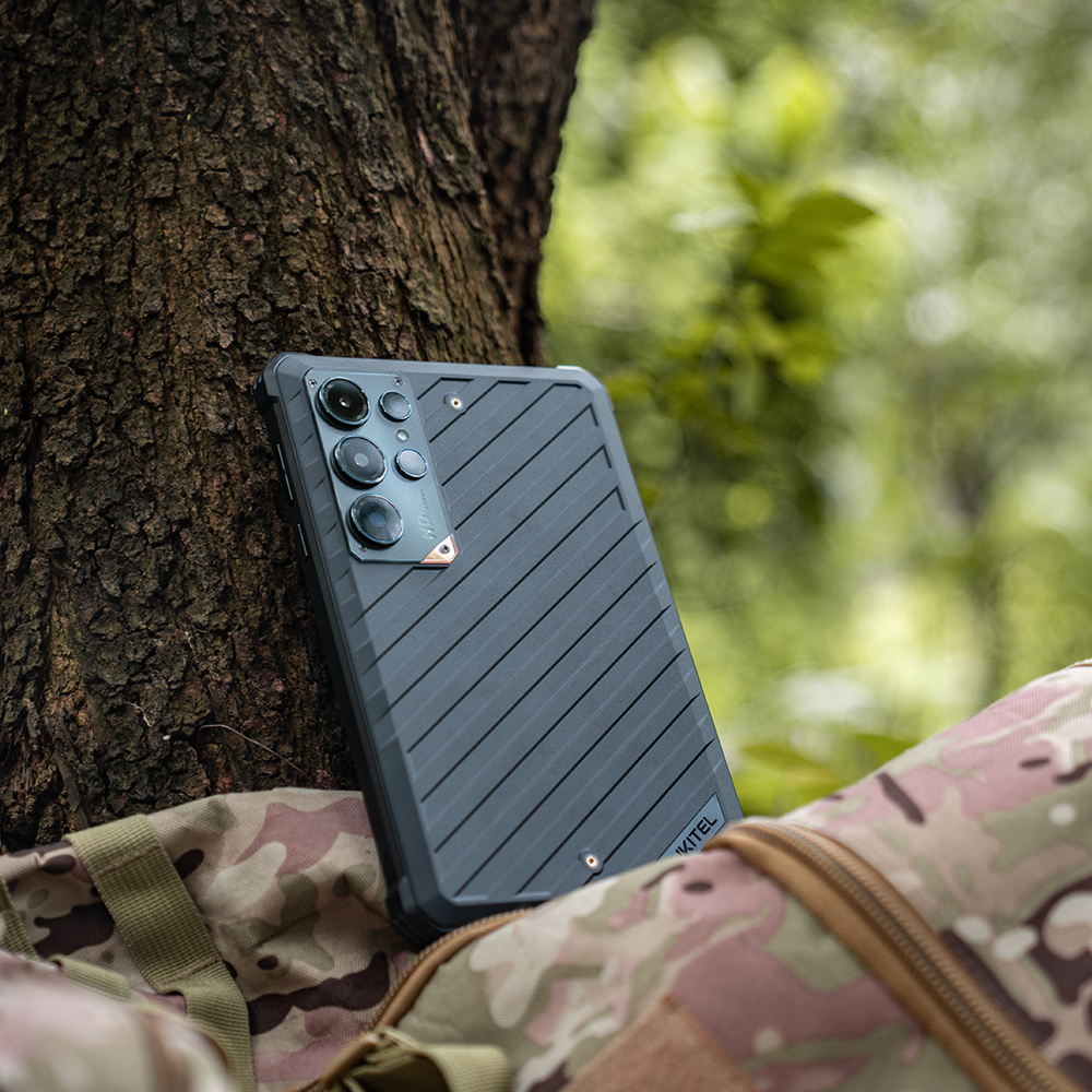 Uno smartphone grigio scuro robusto appoggiato contro il tronco di un albero vicino a uno zaino mimetico.