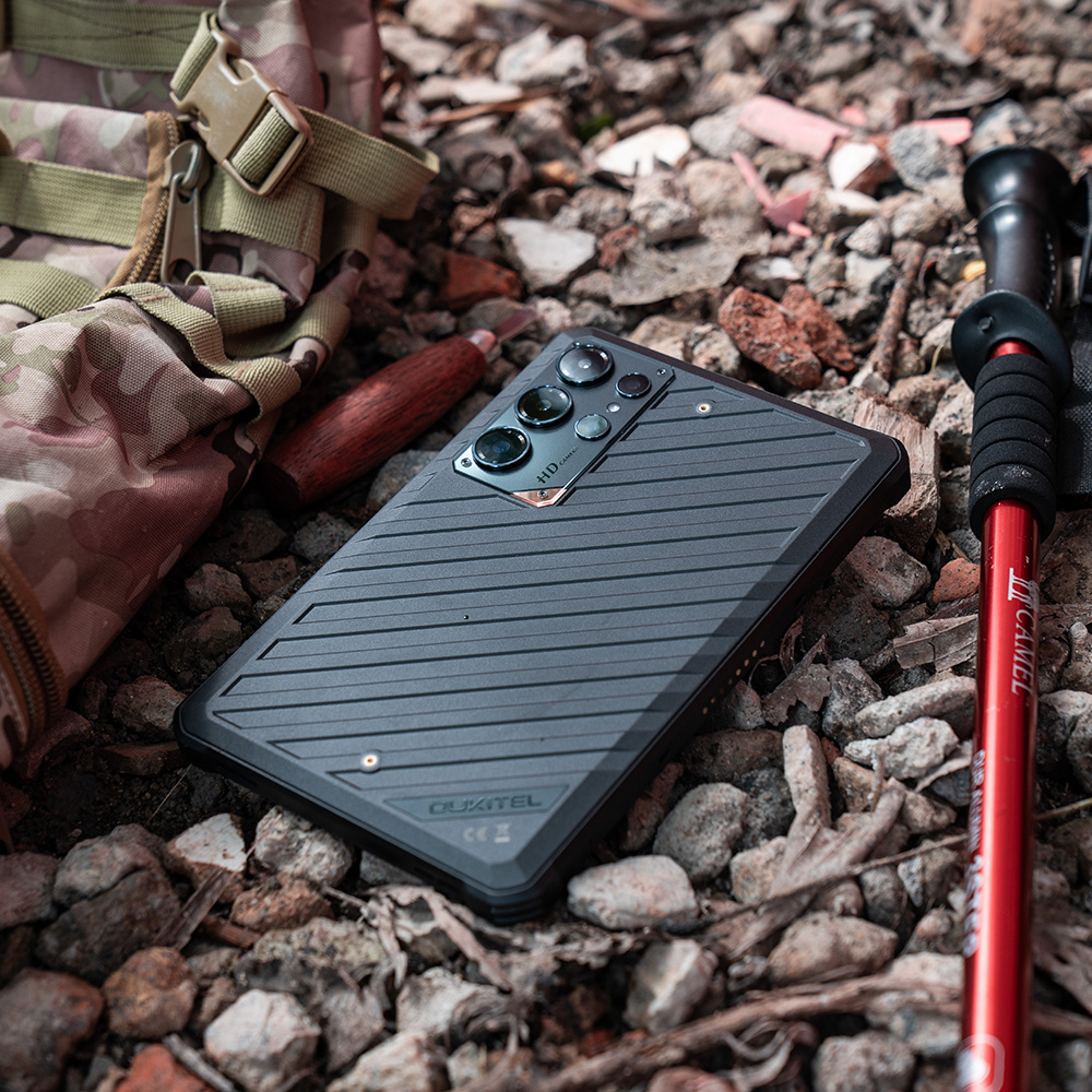 Uno smartphone rugged su terreno roccioso con zaino mimetico e bastone rosso.