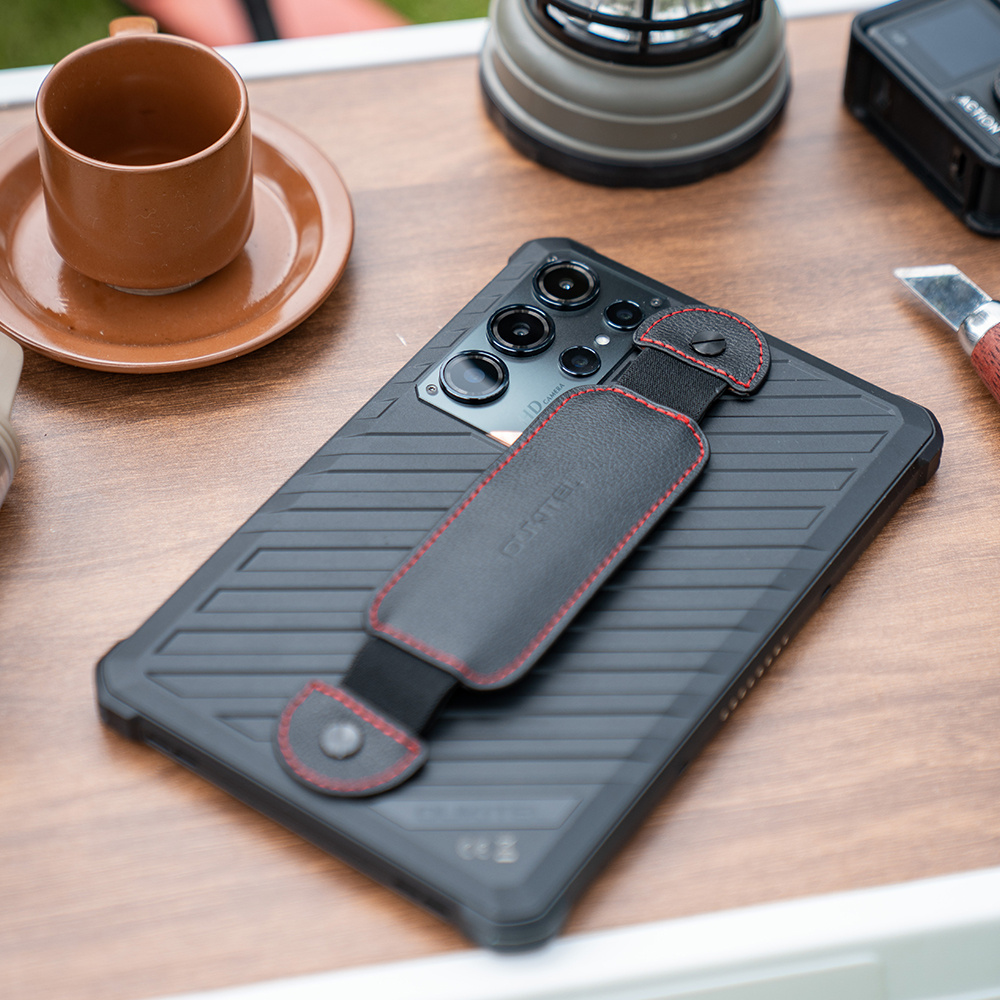Uno smartphone nero con cinturino in pelle su un tavolo di legno, accanto a una tazza marrone.
