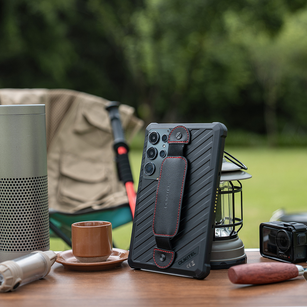 Uno smartphone nero robusto con cinturino in pelle su un tavolo di legno all'aperto, con attrezzatura da campeggio.