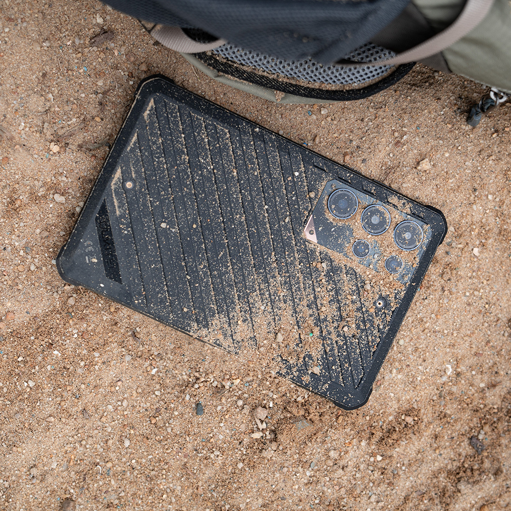Un tablet nero coperto di sabbia con più obiettivi della fotocamera giace su terreno sabbioso.