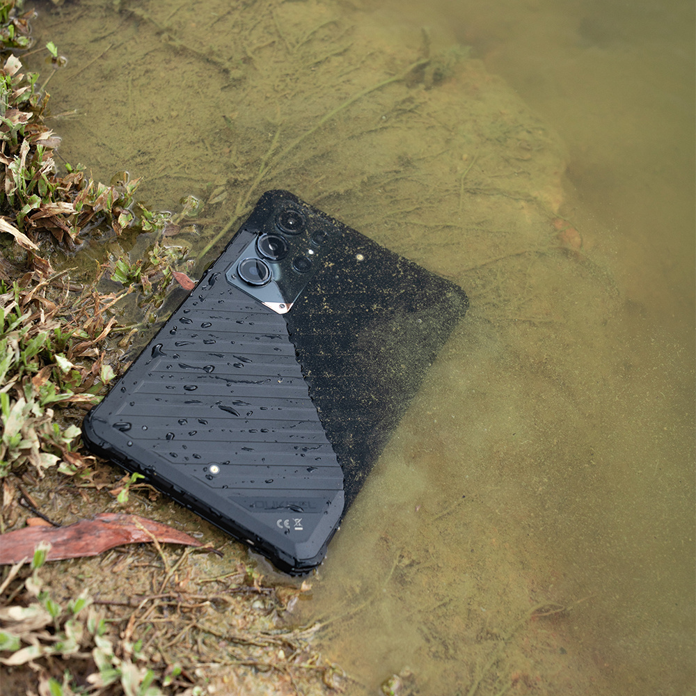 Uno smartphone nero robusto giace in acqua fangosa e poco profonda vicino a una riva.