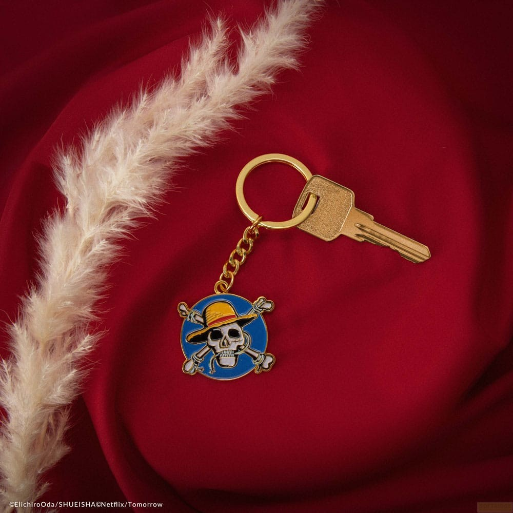 Un llavero dorado con un cráneo y sombrero de paja sobre tela roja junto a una pluma de hierba de la pampa.
