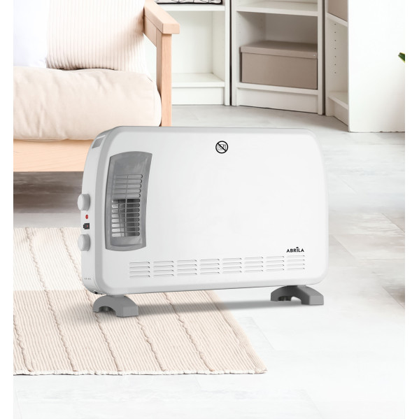 Un calefactor de convección blanco con patas grises y rejilla de ventilación sobre una alfombra en una habitación luminosa.