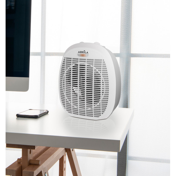 Un calefactor blanco Abrila está en un escritorio frente a una ventana, al lado de un monitor y un teléfono.