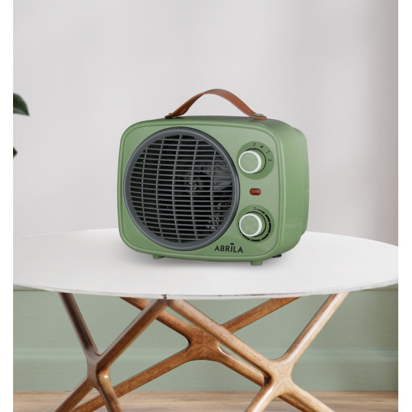 Un calefactor retro verde con asa de cuero está sobre una mesa redonda de madera en una habitación luminosa.