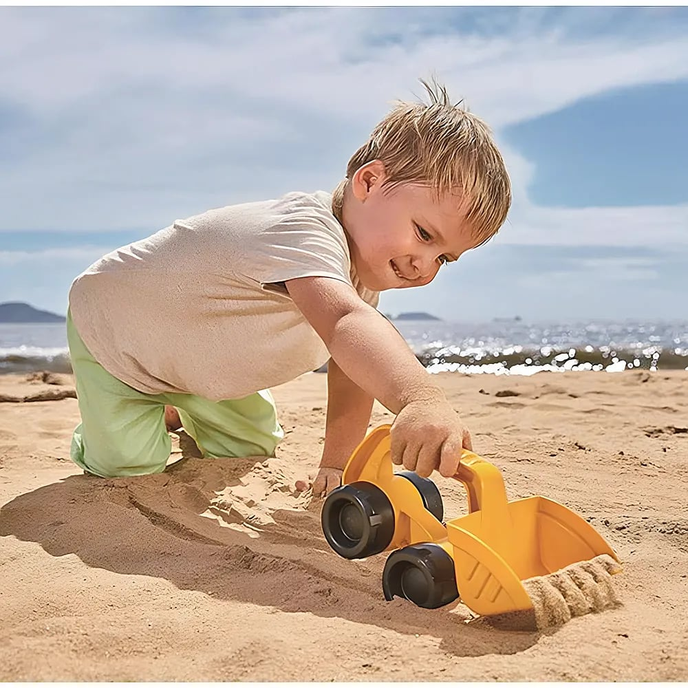 Un enfant genou sur une plage de sable joue avec une pelleteuse jouet jaune, avec la mer scintillante en arrière-plan.