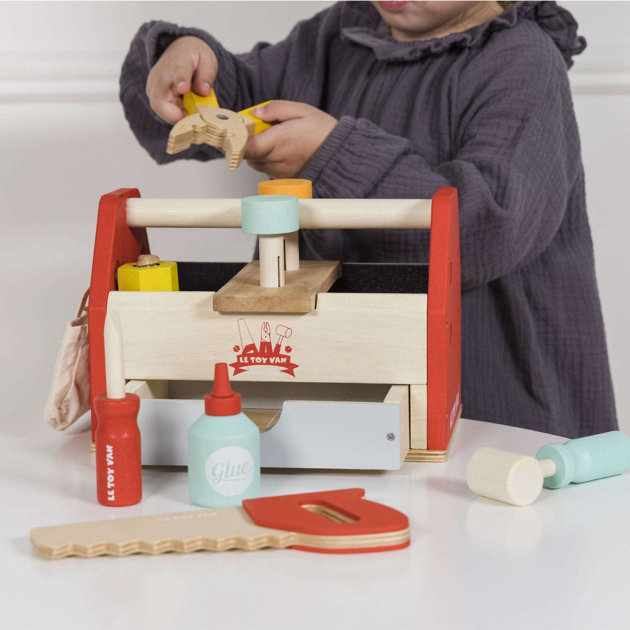 Ein Kind in grauer Kleidung spielt mit einer bunten Holzspielzeug-Werkzeugkiste.