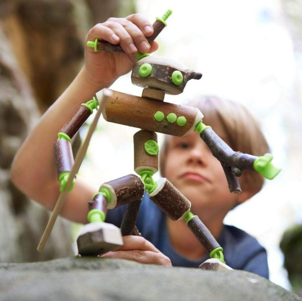 Ein Kind baut draußen eine Spielfigur aus Ästen, die mit grünen Plastikverbindern verbunden sind.
