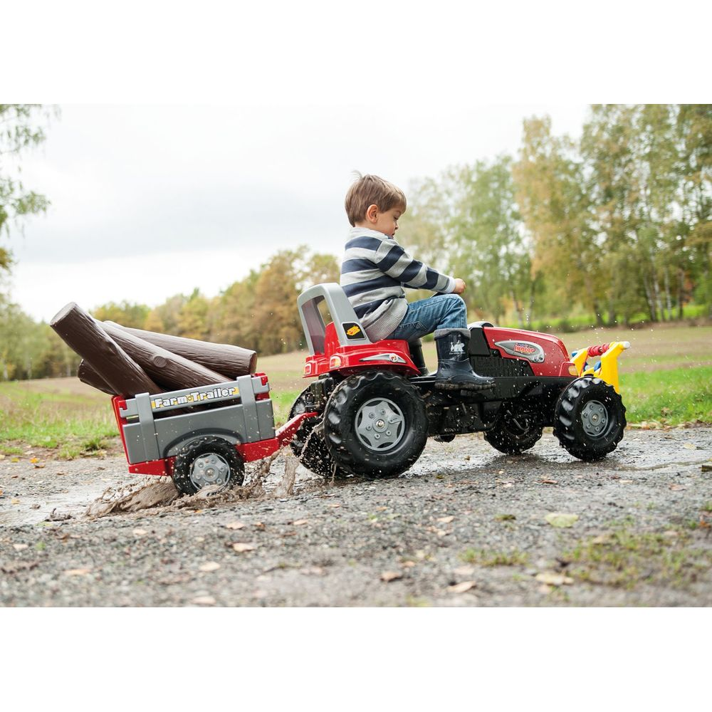Ein Junge fährt einen roten Spielzeugtraktor mit Anhänger und Baumstämmen durch eine Pfütze in der Natur.