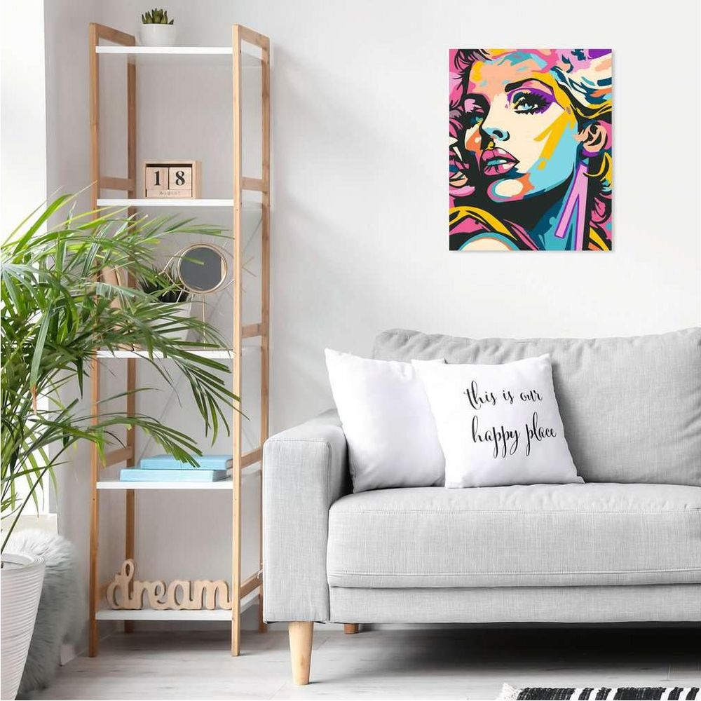 Buntes Pop-Art-Gemälde einer Frau über einem grauen Sofa neben einem Holzregal mit grüner Pflanze.