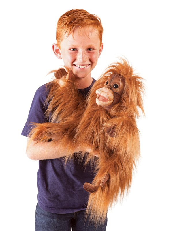 Un garçon roux souriant en t-shirt bleu tient une marionnette orang-outan duveteuse sur fond blanc.