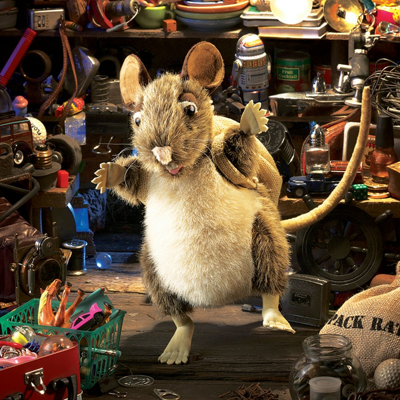Eine flauschige Spielzeugmaus mit Rucksack steht inmitten von Regalen voller Spielzeug und Gegenstände.