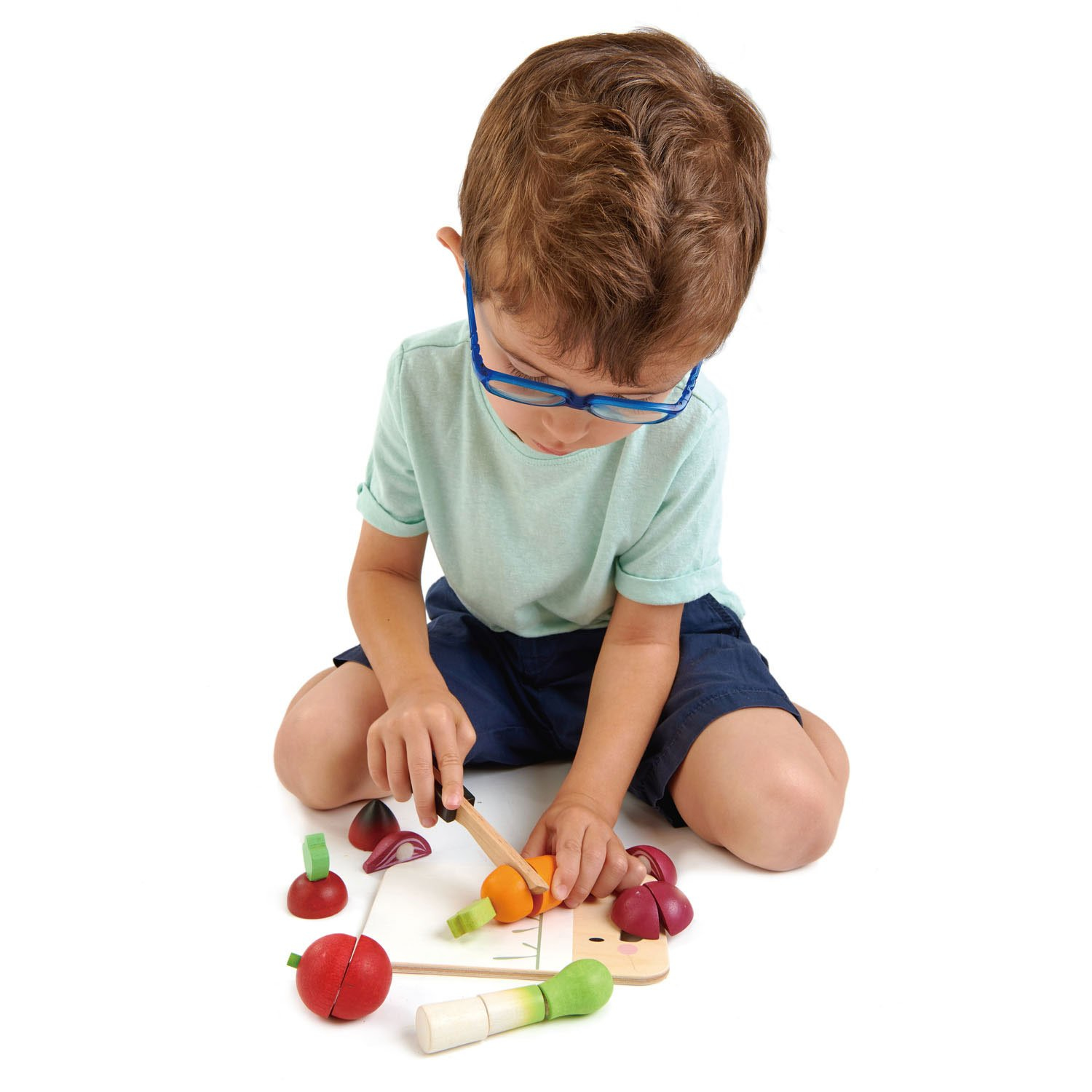 Kleiner Junge mit blauen Brillen schneidet Holzspielzeug-Gemüse auf einem Brett.