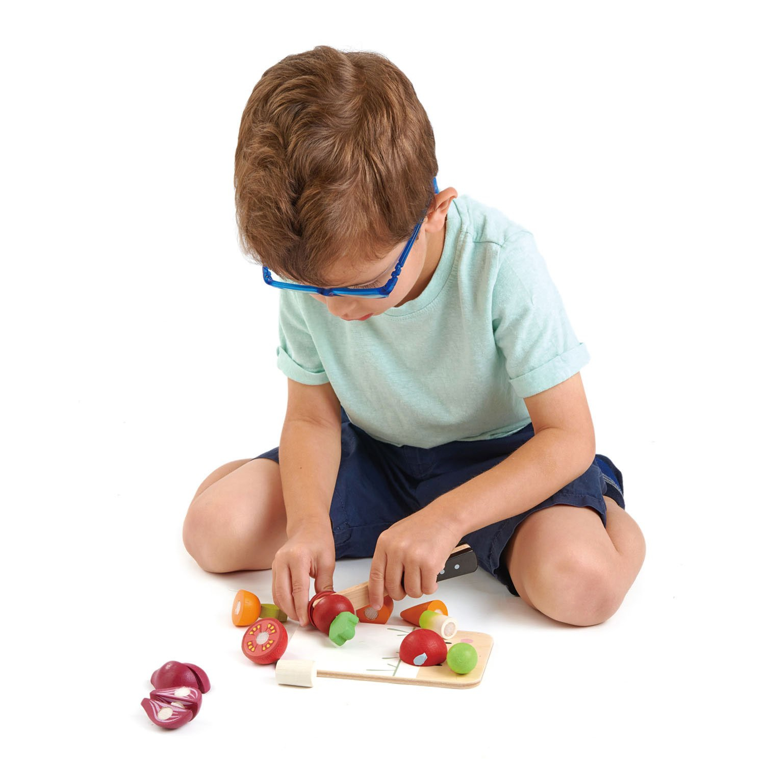 Ein Junge mit blauen Brillen sitzt auf dem Boden und spielt mit Holzspielzeug-Gemüse.