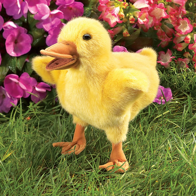 Ein flauschiges gelbes Plüschtier einer Ente im Gras vor rosafarbenen Blumen.