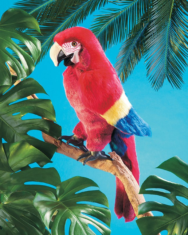 Ein roter Plüschpapagei sitzt auf einem Ast vor tropischen Blättern und blauem Himmel.