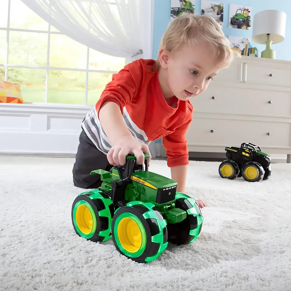 Ein kleiner Junge spielt in einem hellen Raum auf einem weißen Teppich mit einem großen grünen Spielzeugtraktor.