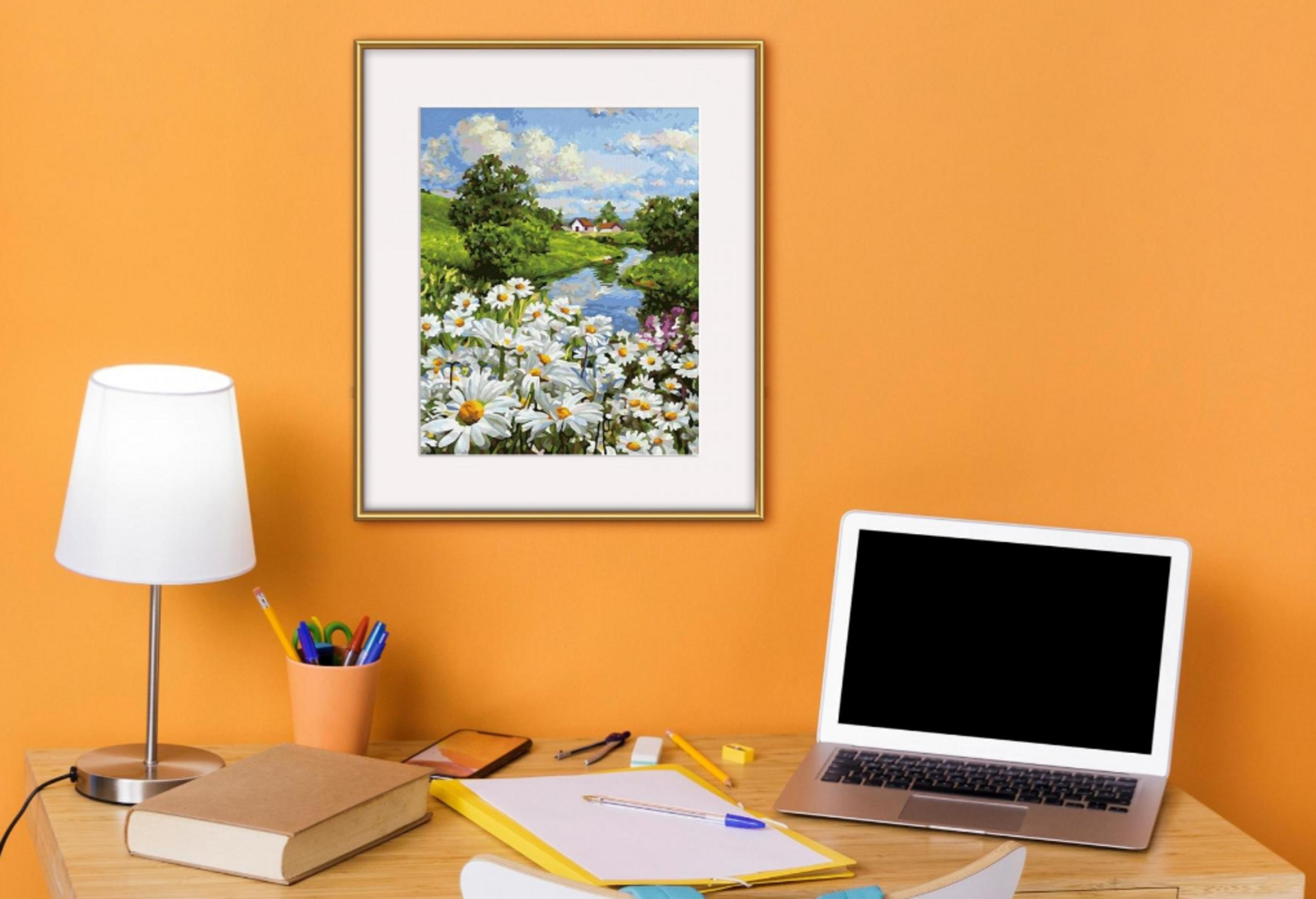 Ein Schreibtisch mit Laptop, Lampe, Buch und einem Landschaftsbild mit Blumen an der orangen Wand.