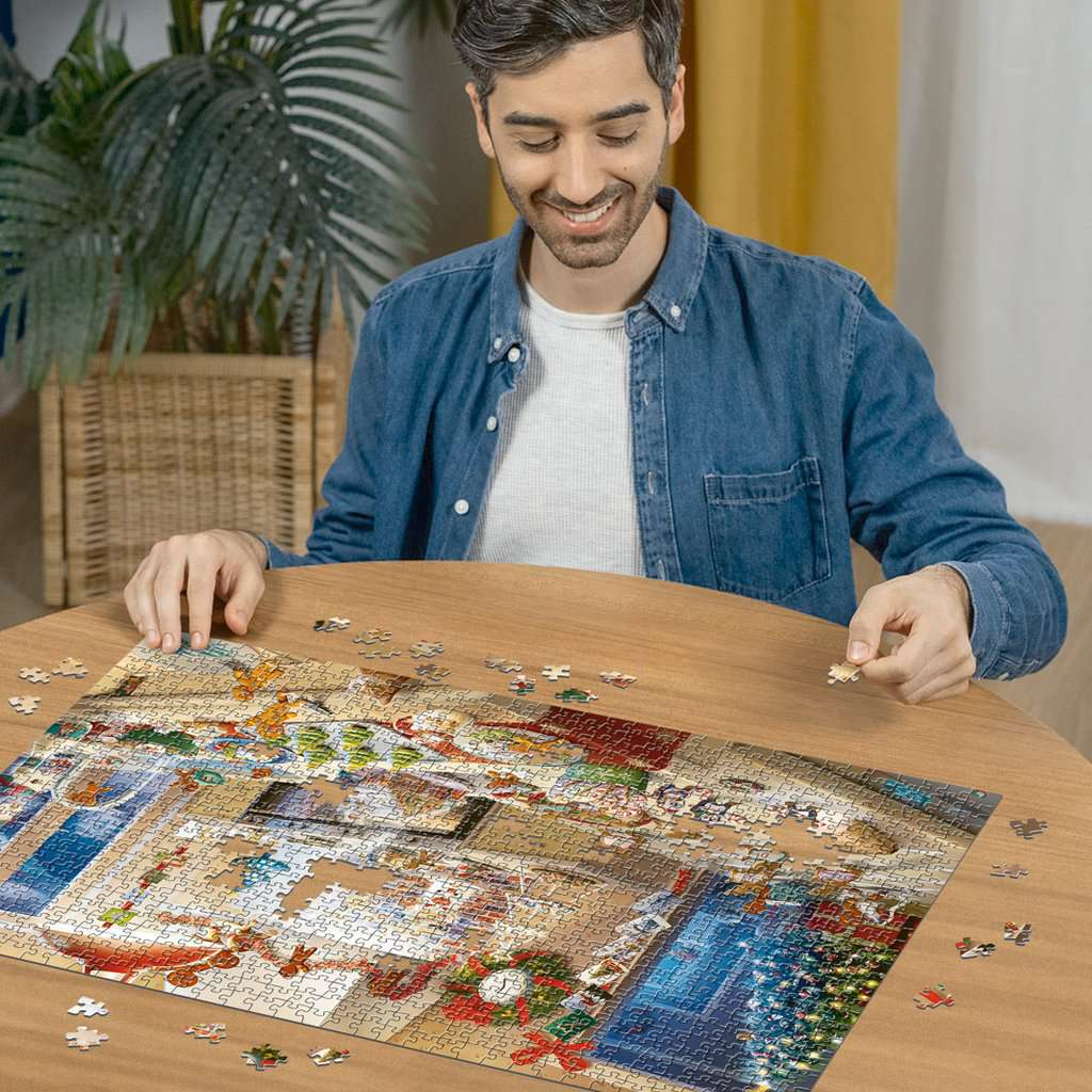 Ein lächelnder Mann in Jeanshemd sitzt an einem Holztisch und löst ein großes Puzzle mit einer Topfpflanze dahinter.