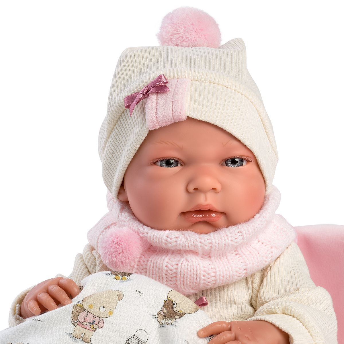 Una bambola con cappello bianco con ponpon rosa, sciarpa rosa e coperta fantasia.