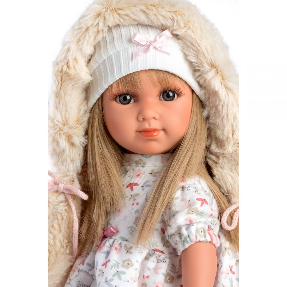 Eine blonde Puppe mit braunen Augen trägt eine Mütze und eine flauschige Fellkapuze.