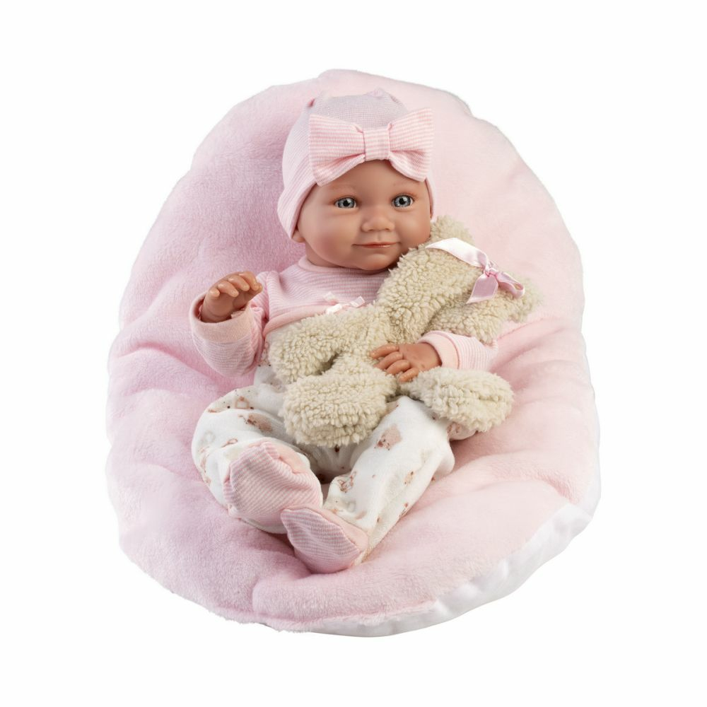 Una bambola realistica con vestiti rosa e cappello siede su un cuscino morbido tenendo un peluche.