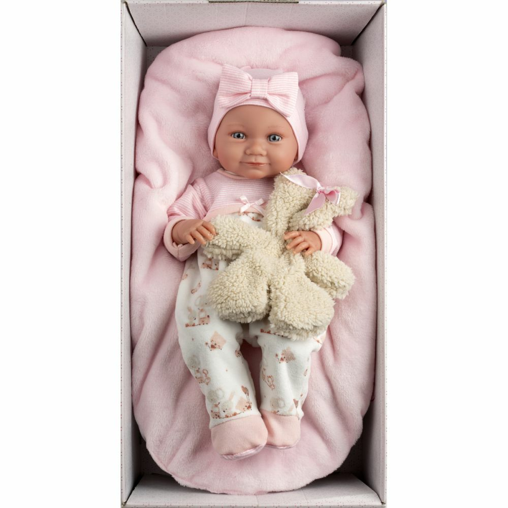 Una bambola con vestiti e cappello rosa giace in una scatola rosa su una coperta, tenendo un peluche.