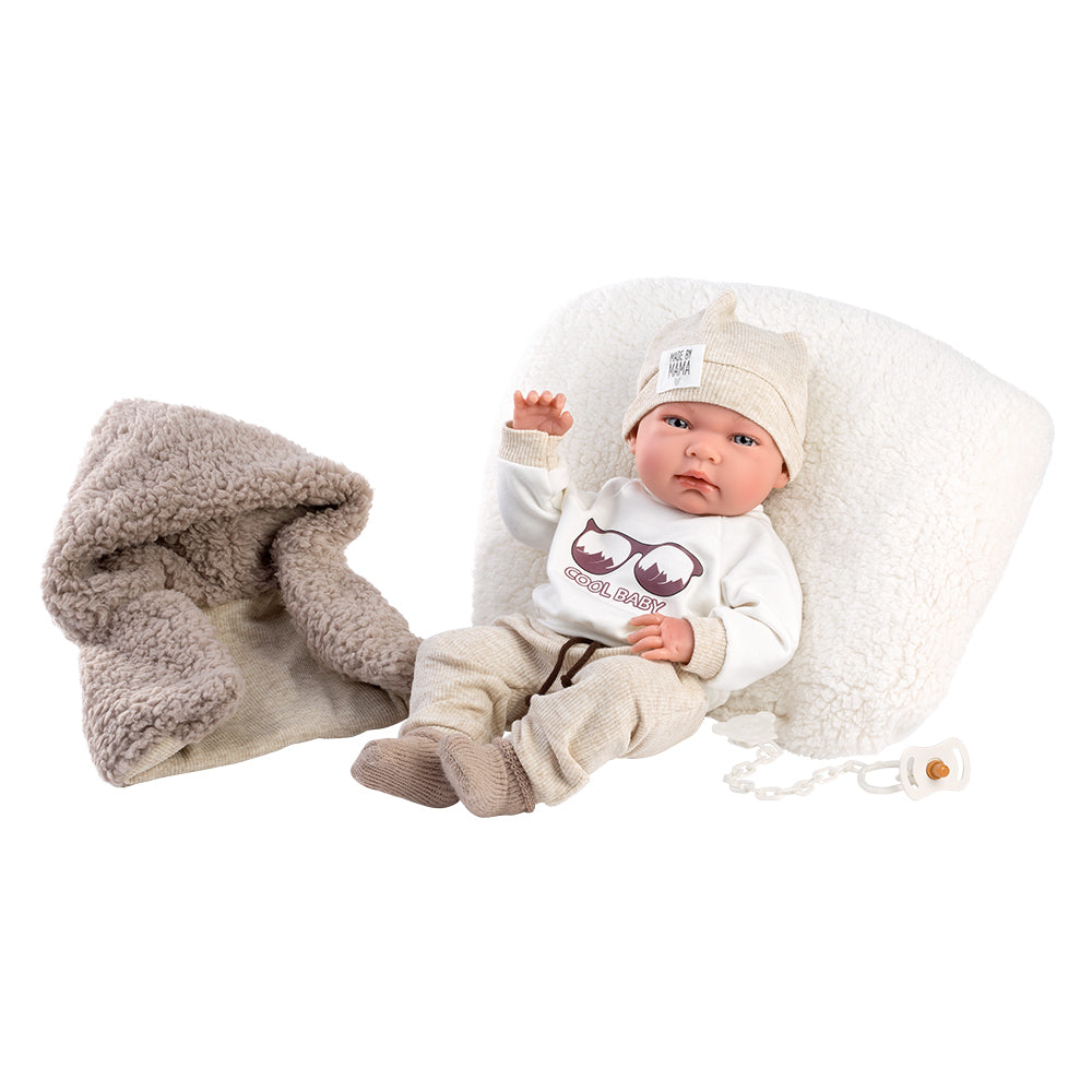 Una bambola con vestiti beige, berretto e ciuccio, riposa davanti a un cuscino soffice e una coperta grigia.